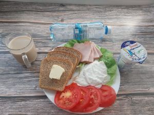 Zdjęcie przedstawia Dietę z ograniczeniem łatwoprzyswajalnych węglowodanów: Masło extra  Kawa zbożowa z mlekiem Chleb Graham  Szynka Piastowska- wp., wędzona, parzona z dod. b.sojowego i wody Jogurt naturalny Woda mineralna Sałata zielona Pasta z twarogu z natką pietruszki Pomidor